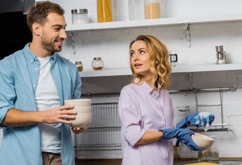 attractive woman in rubber gloves washing dishes and looking at smiling husband holding bowls in kitchen