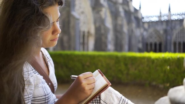 Side view crop young lady with pen and notebook sitting near column inside old abbey in Portugal 