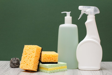 Bottles of dishwashing liquid, glass and tile cleaner and sponges on green background.