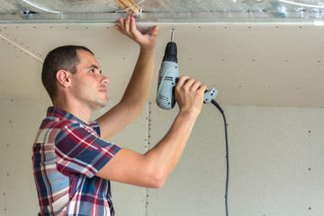Young man in usual clothing and work gloves fixing drywall suspended ceiling to metal frame using...