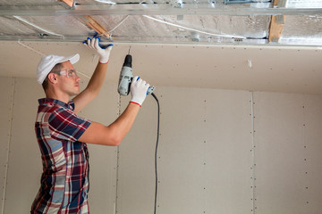 Young man in goggles fixing drywall suspended ceiling to metal frame using electrical screwdriver...