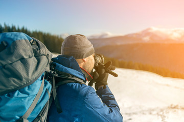 Hiker man tourist photographer in warm clothing with backpack and camera taking picture of snowy...