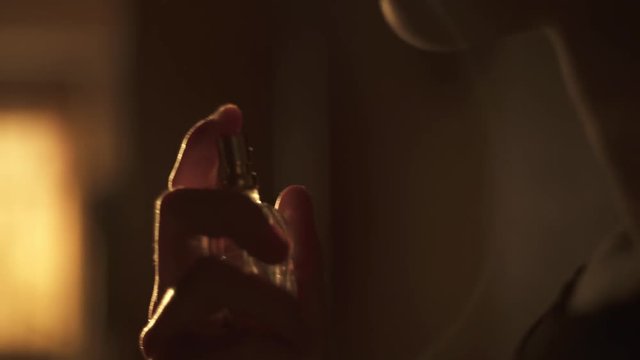 Young woman squirting perfume on her neck. In a room orange sunset light. Splashes fly slowly in the sunlight.