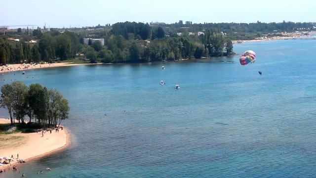 Parasailing by parachute behind boat over waters of blue sea near beach. Man tourist flying on parachute over lake in sunny weather. Vacation parasailing activity