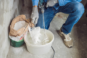 Young male painter kneads putty with water in a bucket using a hand-held mixer for building mixes
