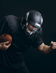 American football caucasian player wearing black uniform with protection holding ball isolated over black studio wall