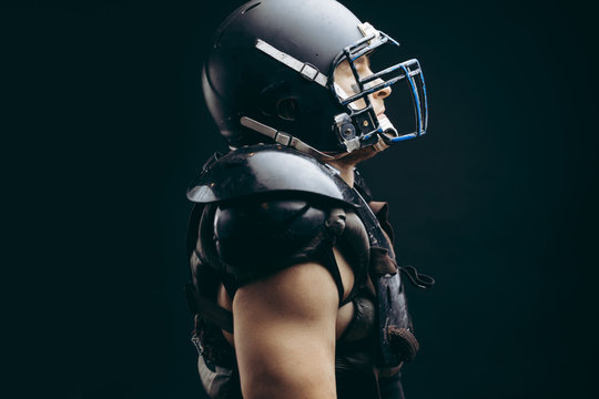 Head and shoulder portrait of muscular American football player wearing black helmet and protective armour over naked torso, looking at camera over black background
