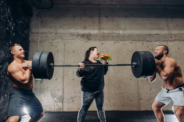 yummy hamburger destroys training. bossy plump woman with fast food is making sportsmen working out with huge barbell