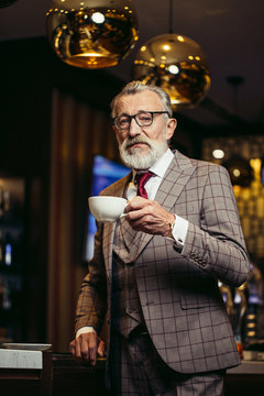 Senior businessman in rich formal three piece suit drinking coffee standing near bar counter at dark restaurant interior and looking at camera