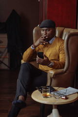 African american sales manager male in yellow coat and black hat waiting for his client in restaurant lounge, sitting in arm chair, spending time browsing internet on smartphone