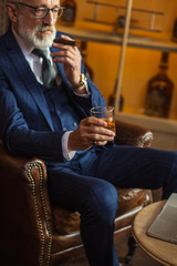 Well-dressed experienced caucasian old-aged CEO relaxing with cigar and alcohol, reading news on...