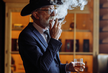 Portrait of old-aged male dandy dressed in elegant trendy men s suit in big hat smokes cigar and drinks alcoholic beverage.