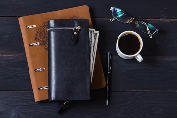 Men's wallet, notebook, cup of coffee and glasses on a black wooden table. View from above.