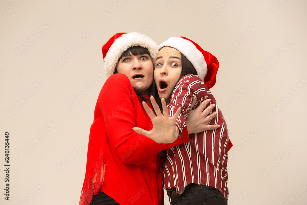 Poster A portrait of a scared mother and daughter in Santa hat at studio on gray background. Trendy colors. Human positive emotions and facial expressions concept