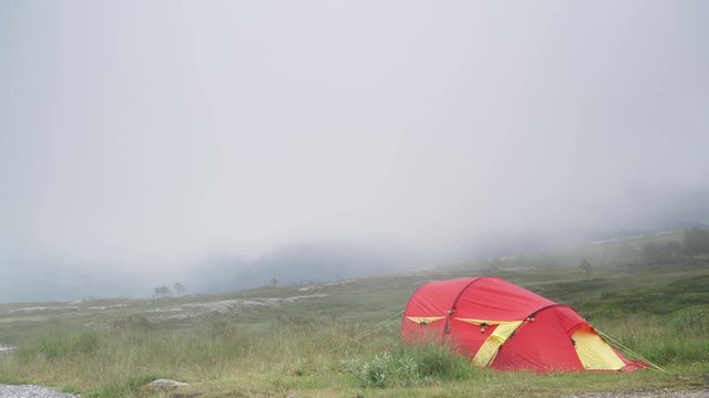 Red tent in norwegian mountains with moving rain clouds. Rainy windy weather. Camping on nature. Holidays and travel