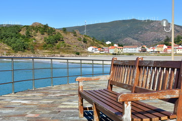 Wooden bench on a beach promenade. Turquoise water and small coastal village. Sunny day, Galicia, Spain.