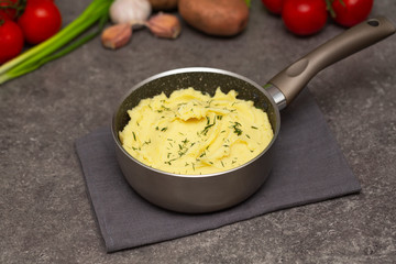 Mashed potatoes in a saucepan on dark background, top view. Delicious hot mashed potato with butter and finely chopped fresh herbs. Boiled puree in a saucepan on dark background, top view