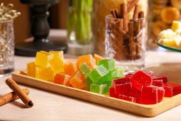Closeup of stacks of small cubes of green, red, yellow, orange marmalade and cinnamon sticks in crystal glass on rectangular wooden plate on catering table