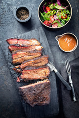 Traditional smoked barbecue wagyu beef brisket offered as top view on an old cutting board with Louisiana sauce and salad with tomatoes