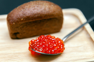 Small piece of black bread with cumin and metal spoon with slide of red salmon caviar gourmet snack on rectangular wooden plate on black background. Concept healthy seafood appetizer