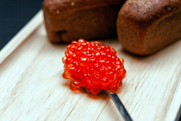 Closeup macro two small pieces black bread with cumin and metal spoon with slide of red salmon caviar gourmet snack on rectangular wooden plate on black background. Concept healthy seafood appetizer
