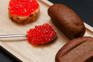 Closeup small pieces black bread with cumin, metal spoon with slide of red salmon caviar, sandwich on rectangular wooden plate black background. Concept gourmet snack healthy seafood appetizer