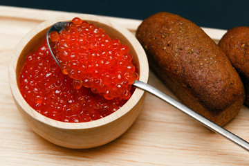Closeup macro small buns black bread with cumin, saucer slide red salmon caviar and metal spoon on rectangular wooden plate on black background. Concept gourmet snack healthy seafood appetizer