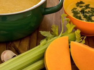 Pumpkin and carrot soup with parsley in bowl and sousepan on dark wooden table with selery, garlick...