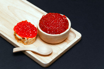 Closeup macro small red salmon caviar canape sandwich on white bread, saucer with slide, little spoon on rectangular wooden plate on black background. Concept gourmet snack seafood appetizer