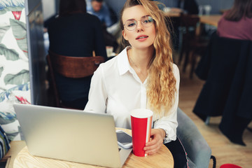 Woman Wear Glasses, Drink Her Hot Coffee While Work In Cafe On Her Laptop. Portrait Of Stylish Smiling Woman In Winter Clothes Drinking Hot Coffee And Work At Laptop. Female Winter Style. - Image