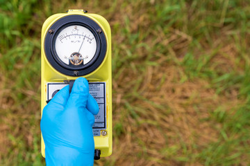 A blue gloved hand holds a radiation meter. The meter shows a high level of radiation. Grass in the...