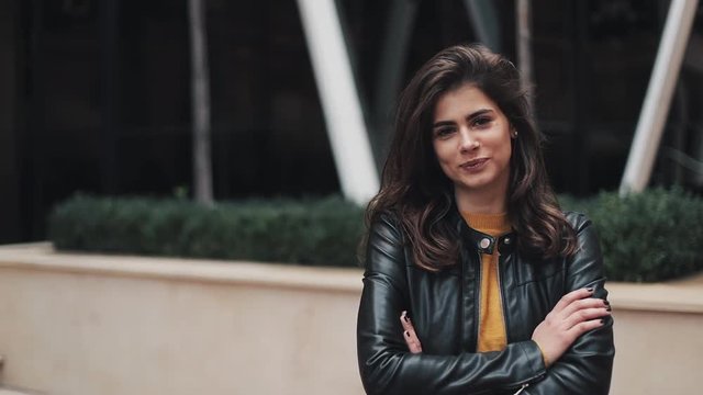 Portrait shot of the young attractive woman standing near office building smiling to the camera with hands crossed in front of her. She is dressed in a warm yellow sweater and autumn leather jacket