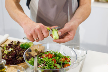 Chef cutting cucumber for salad in light kitchen. Preparing fresh salad in glass bowl. Cooking in cozy cuisine at home. Family dinner on holidays