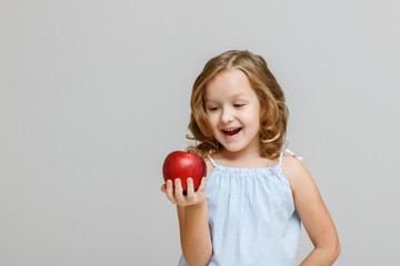 Portrait of a happy smiling little blonde girl on a gray background. The child is looking and ready to eat red apple