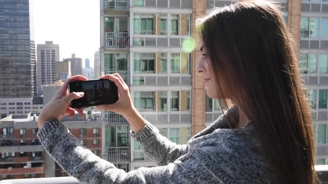 Young millennial beautiful woman or tourist taking photograph with mobile cellphone smartphone device of skyline in New York view from her apartment. Girl to share pic on social media photo site.