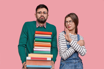 Photo of busy bearded male student carries many books, displeased female shows disagreement gesture, refuses to help with exam preparation, model together against pink background. Studying concept