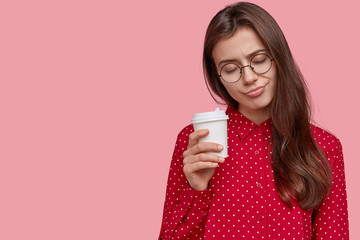 Overworked dark haired woman has sleepy look, carries takeaway coffee in hands, naps during break, dressed in stylish outfit, models over pink studio wall, copy space for your advertising content