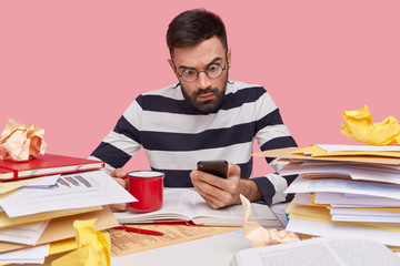 Indoor shot of stunned emotional young unshaven man holds modern cellular, wears transparent round glasses and striped jumper, drinks aromatic hot coffee, surrounded with many papers, notepad