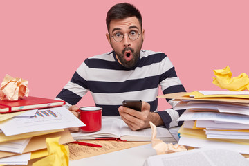 Indoor shot of shocked amazed young Caucasian man uses modern cell phone, drinks coffee, writes records in notepad, studies papers with graphics, analyzes information and data, surfes internet.