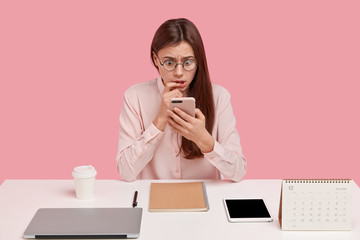 Photo of stupefied European woman browses internet on modern cell phone, has scared expression,...