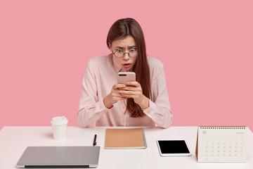 Surprised Caucasian young woman with bated breath, uses new smartphone for chatting and...