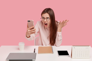 Photo of outraged annoyed woman holds modern cell phone, makes video call, argues with colleague,...