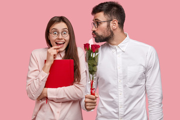 Happy female student in spectacles, carries red textbook, glad to recieve bouquet of roses from male wonk, congratulates her with birthday, isolated over pink background. Two nerds have date together