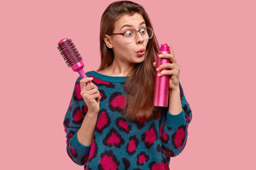 Attractive female wears big square glasses, stares with stupefaction at hairspray, holds hairbrush,...
