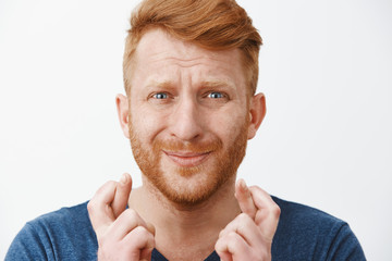 Studio shot of worried intense redhead miserable guy with beard in blue t-shirt crossing fingers, pursing lips and frowning while feeling nervous and begging for good luck and fulfillment of wish