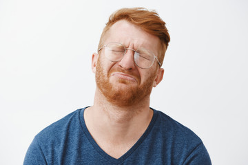 Close-up shot of cute and funny adult with red hair and beard, crying and being gloomy, closing eyes, frowning, tilting head back while pursing lips, feeling regret or upset over gray background