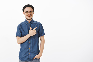 Carefree happy and satisfied handsome male photographer in black trendy beanie and blue shirt pointing at upper right corner smiling broadly with satisfaction and delight over gray background