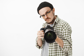 Obraz premium Waist-up shot of funny serious-looking male photographer in black beanie glasses and checked shirt pointing professional camera forward and gazing seriously to take picture during work