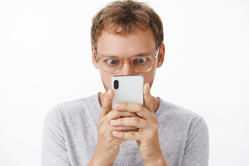 Close-up shot of amused and entertained male geek in glasses holding smartphone close to face...