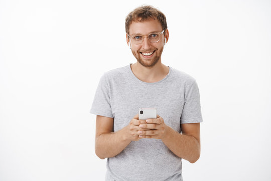 Waist-up shot of satisfied happy male customer with bristle in glasses and grey t-shirt holding brand new smartphone wearing wireless earphones delighted with good quality of sound